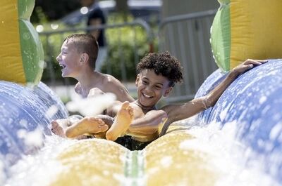 Twee kinderen glijden lachend van een waterglijbaan, omgeven door spuitend water en een kleurrijke achtergrond.