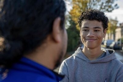 Twee jongeren staan in gesprek, terwijl de zon schijnt en bomen op de achtergrond zichtbaar zijn.