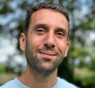 Portret van een man in een blauw T-shirt, glimlachend met een groene, natuurlijke achtergrond.